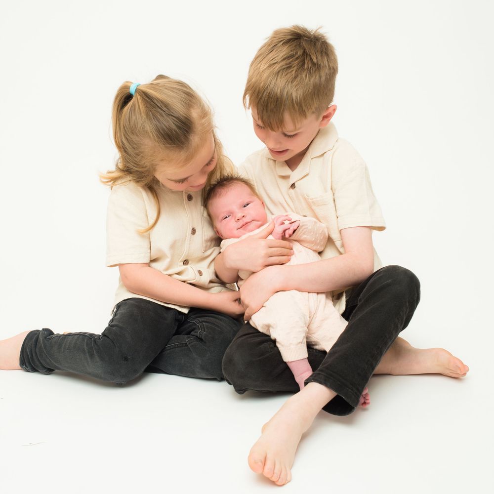 Sibling gently kissing their baby brother during a  newborn photo session  by Abigail's Studio