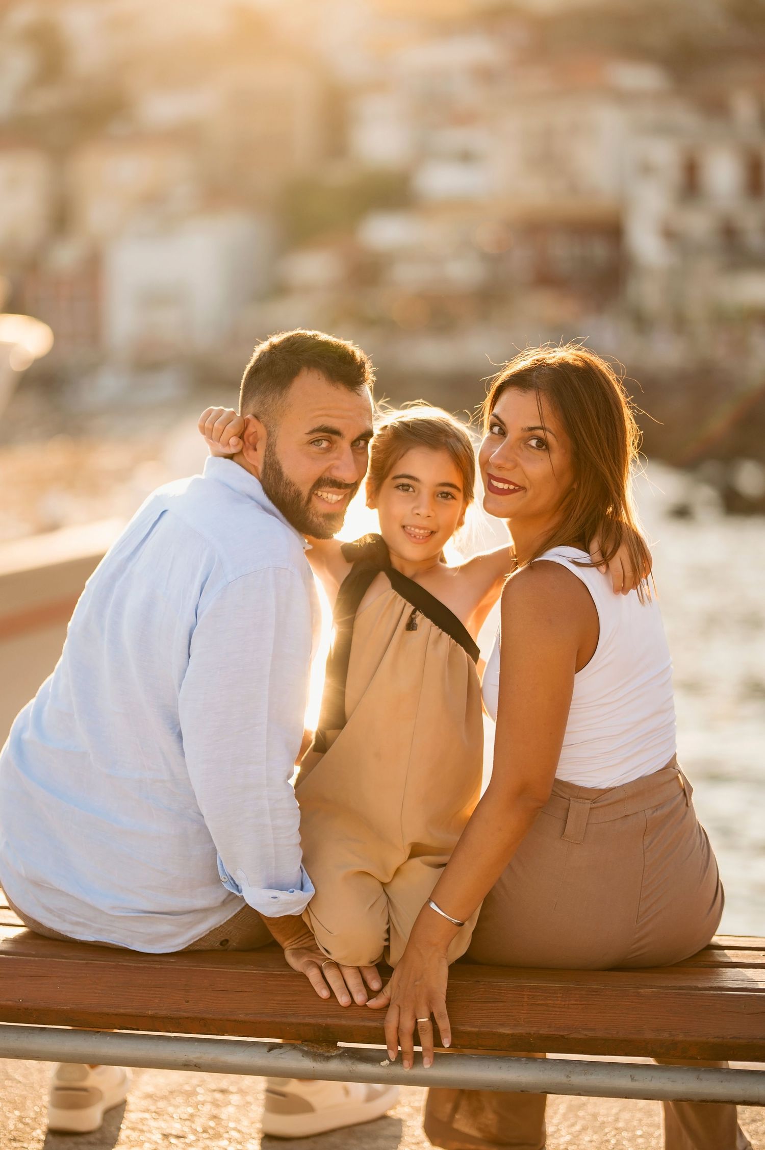 Three people sit together on wooden bench during golden hour sunset with cityscape in background. Parga, Greece