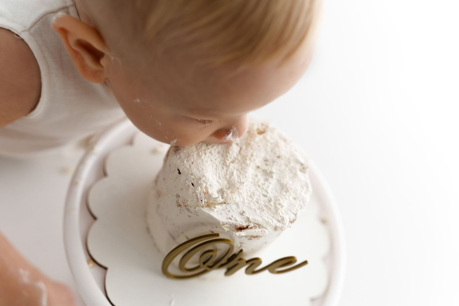Small child bending down to examine a white frosted smash cake with 'One' written on the plate.