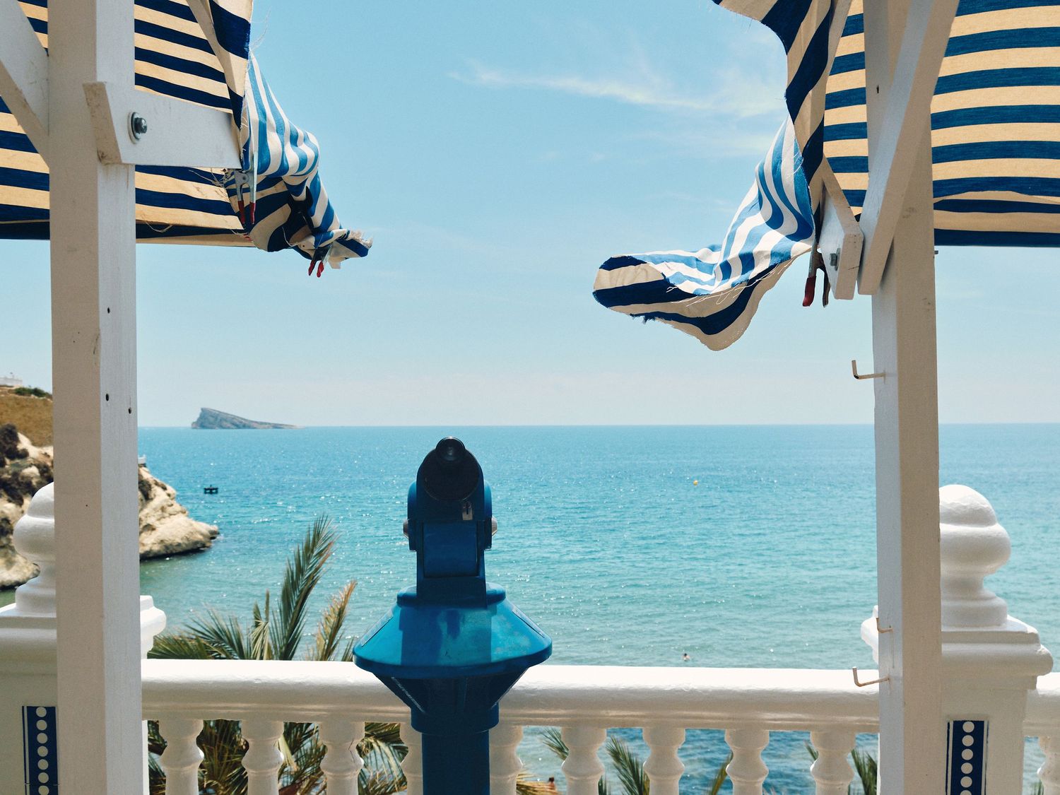 Scenic ocean view from white balcony with blue and white striped awnings overlooking turquoise Mediterranean waters.
