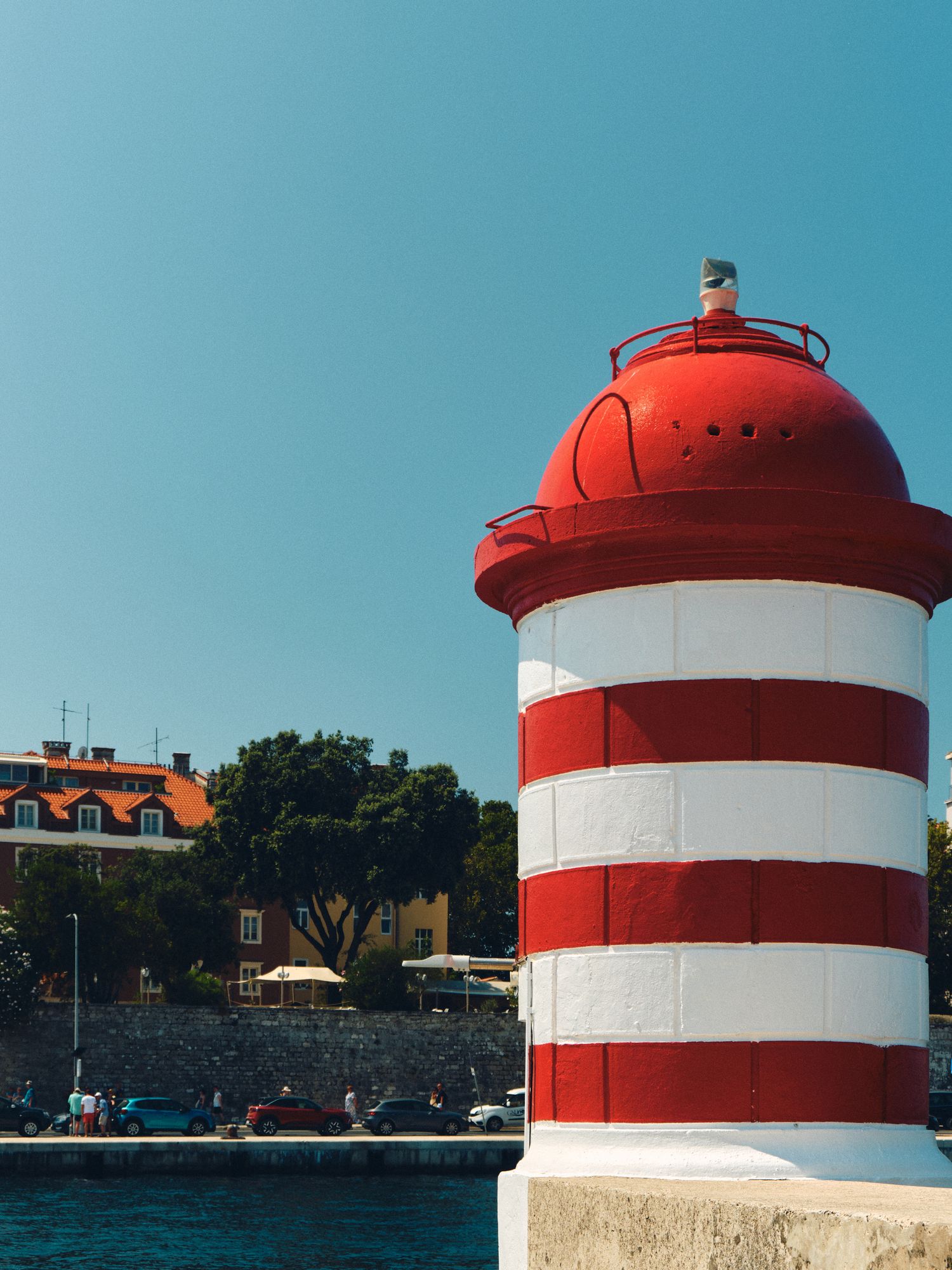 Red and white striped lighthouse tower against clear blue sky with coastal buildings in background.