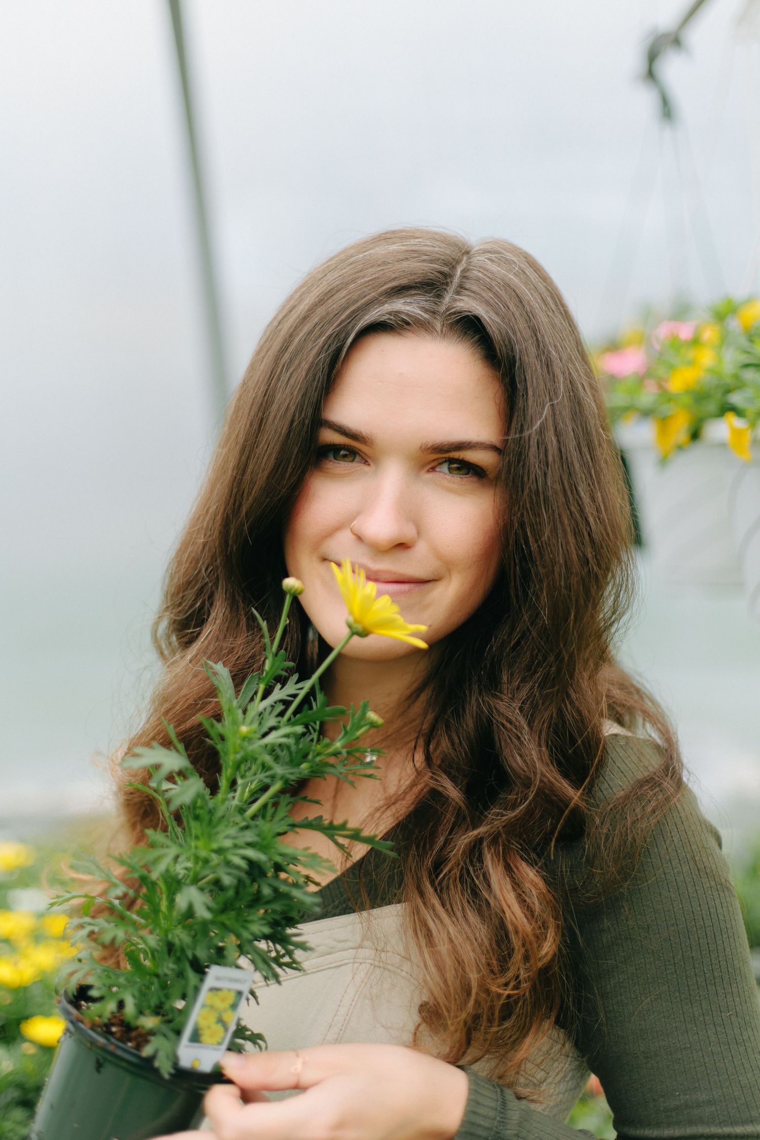 Young woman with long brown hair and green sweater holds potted flowers in a greenhouse setting.