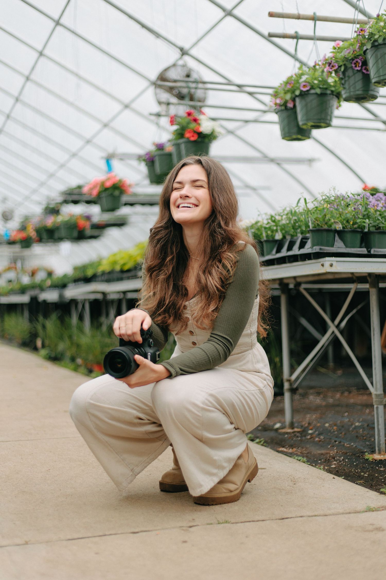Photographer crouching with camera in greenhouse surrounded by hanging flower baskets and plants.