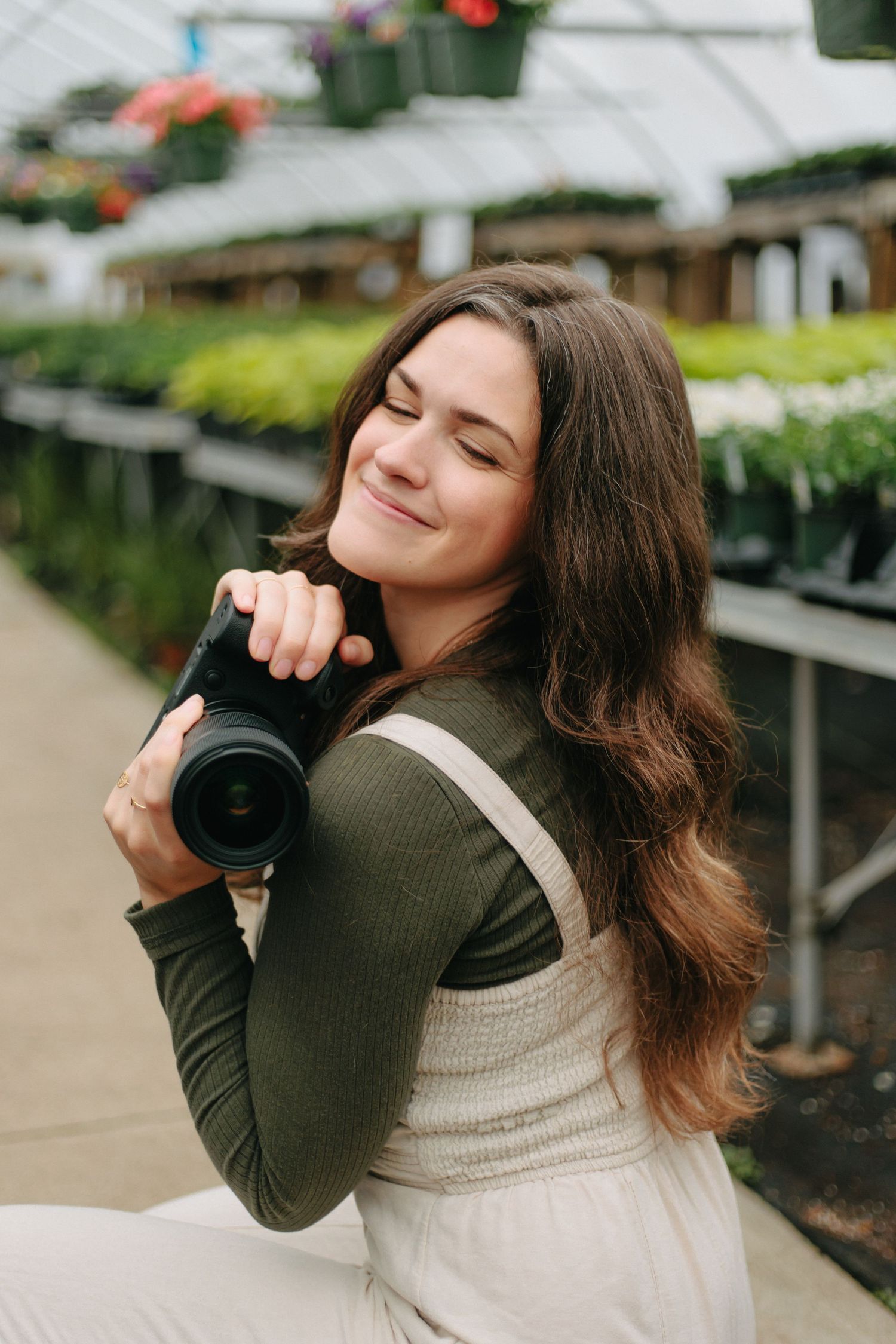 A photographer in a casual dress and olive cardigan smiles while holding a camera in a greenhouse setting.