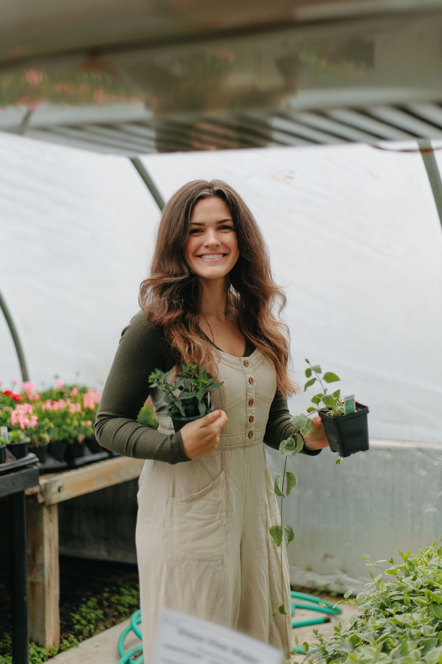 A person wearing a dress and cardigan tends to plants inside a greenhouse filled with flowers and greenery.