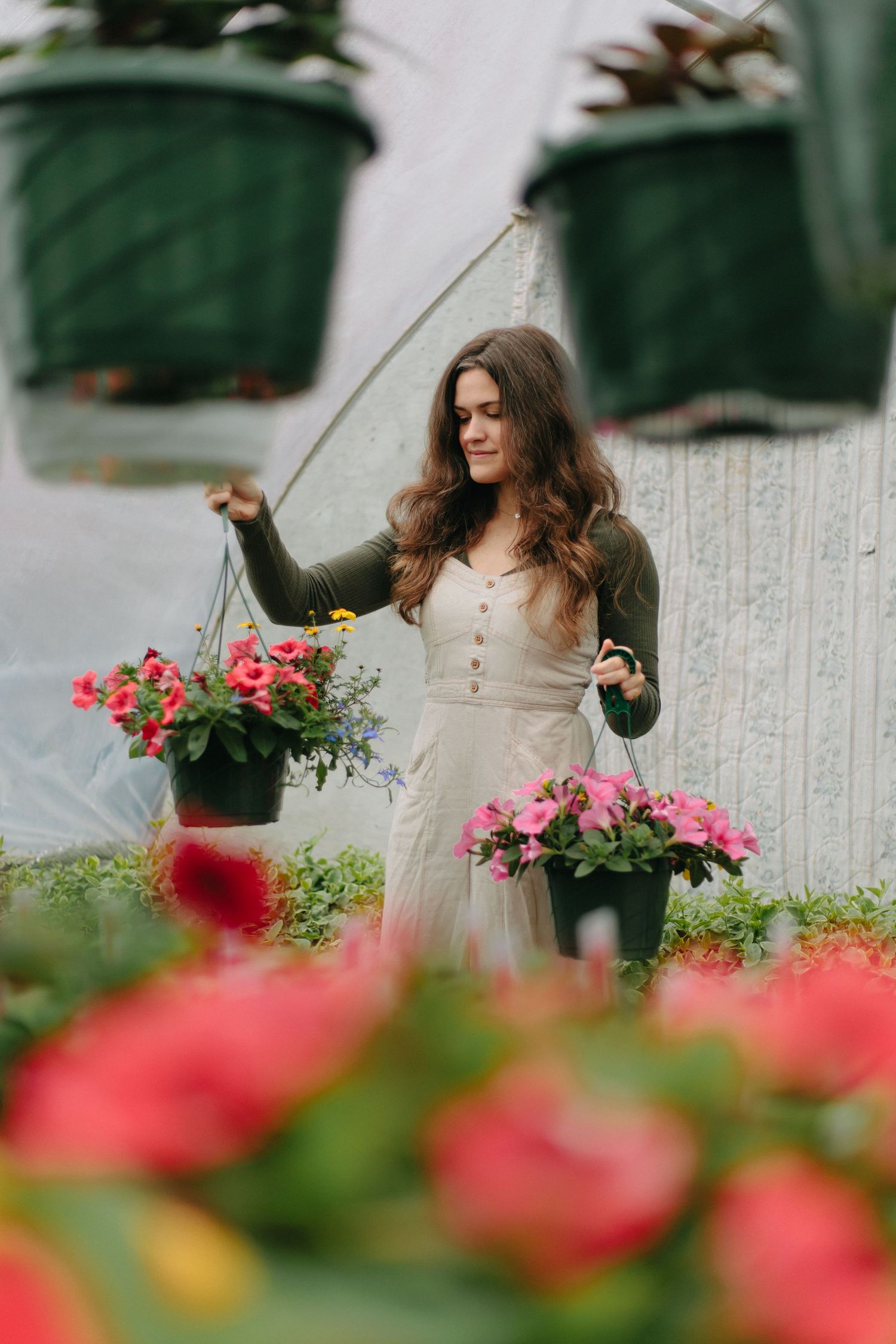 Long-haired gardener in cream dress and black cardigan tends to hanging flower baskets with pink blooms.