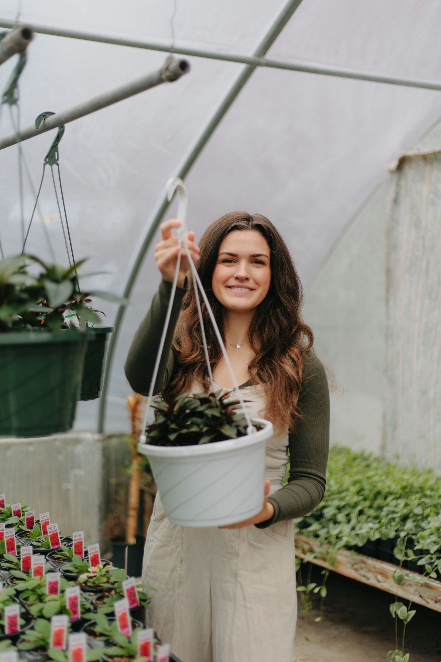 A person in an olive green top holds a white hanging planter inside a greenhouse with potted plants.
