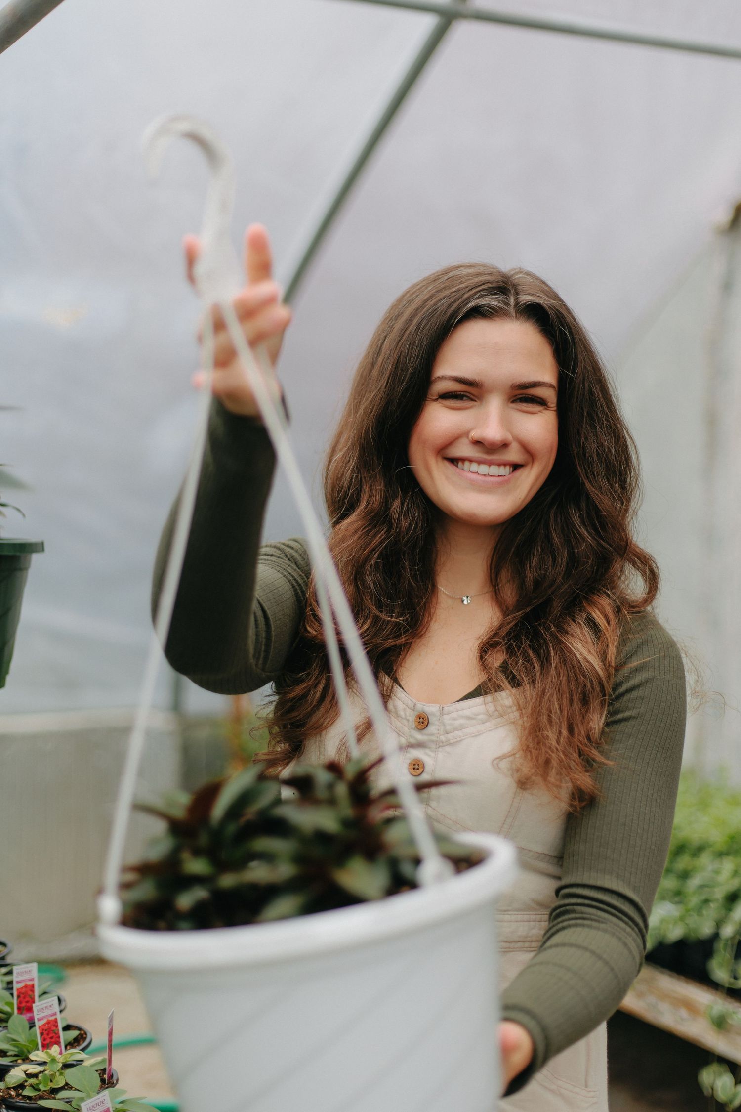 Someone in an olive green sweater and white top tends to hanging plants in a bright greenhouse.