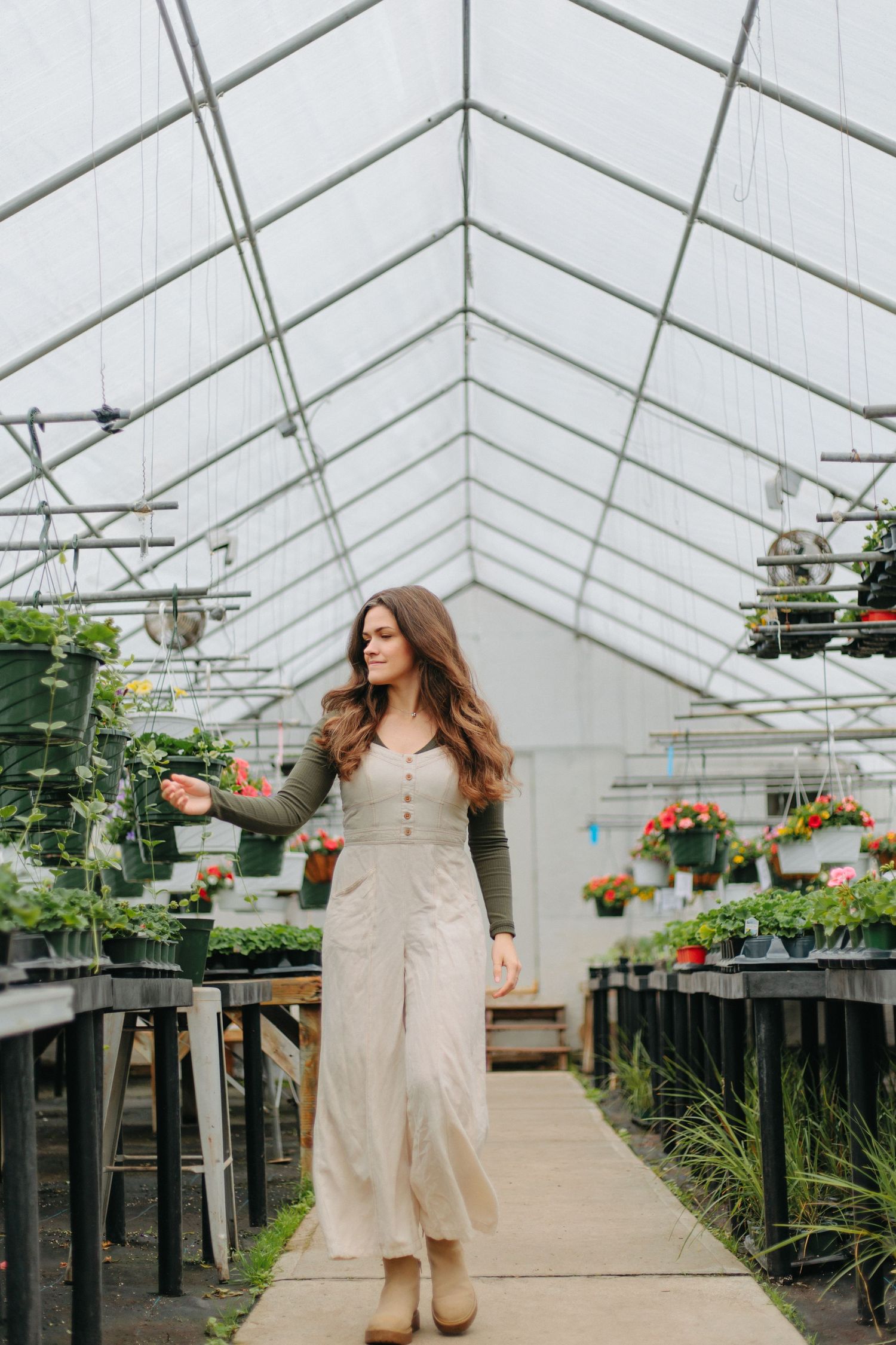 Woman in a beige dress walks through greenhouse filled with potted plants and flowers on elevated tables.