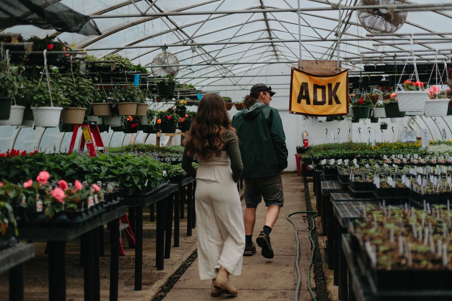 Two people walking through a greenhouse filled with potted plants and hanging baskets under a clear roof.