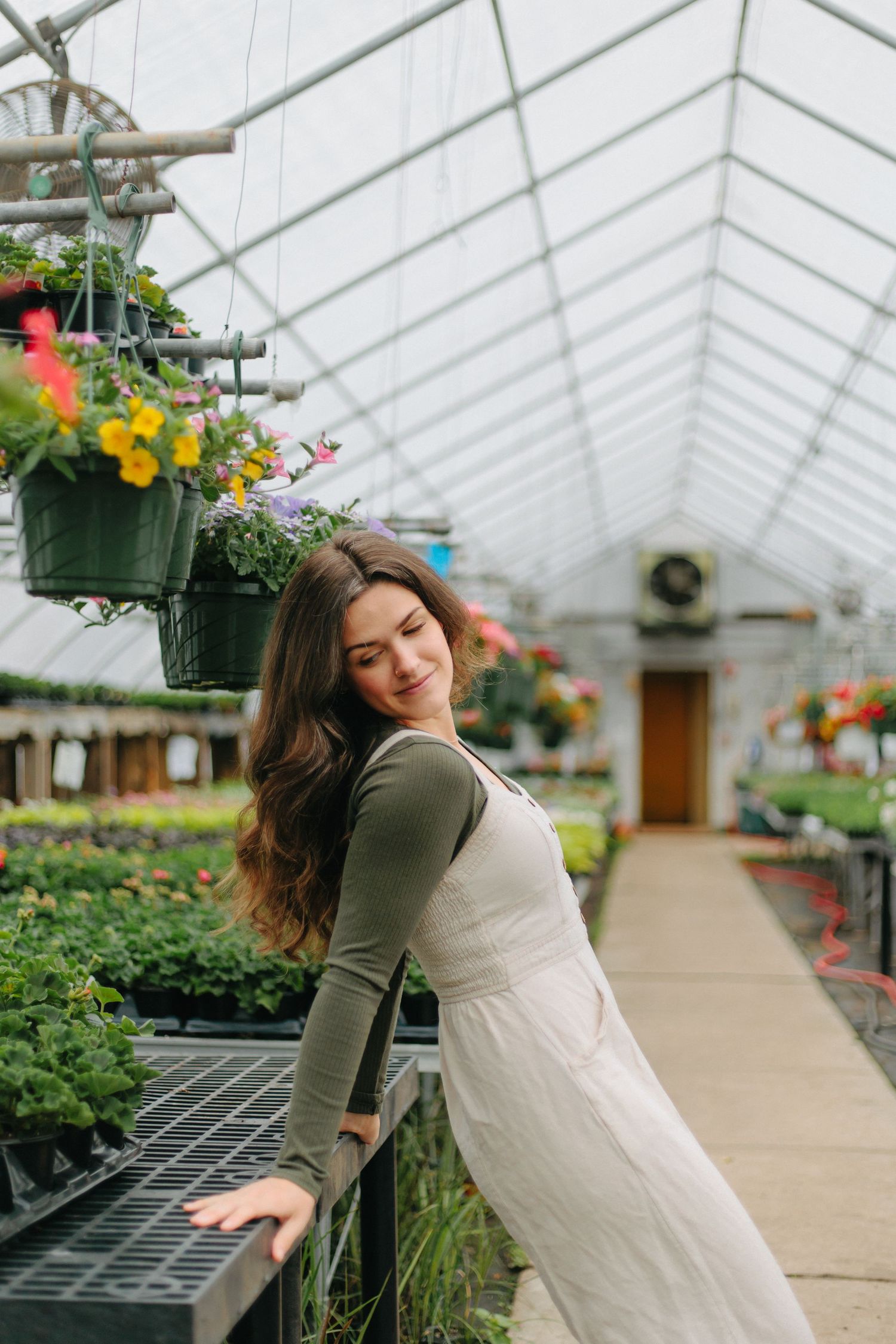 A person in a white dress and green cardigan poses gracefully in a greenhouse filled with colorful hanging flowers and plants.
