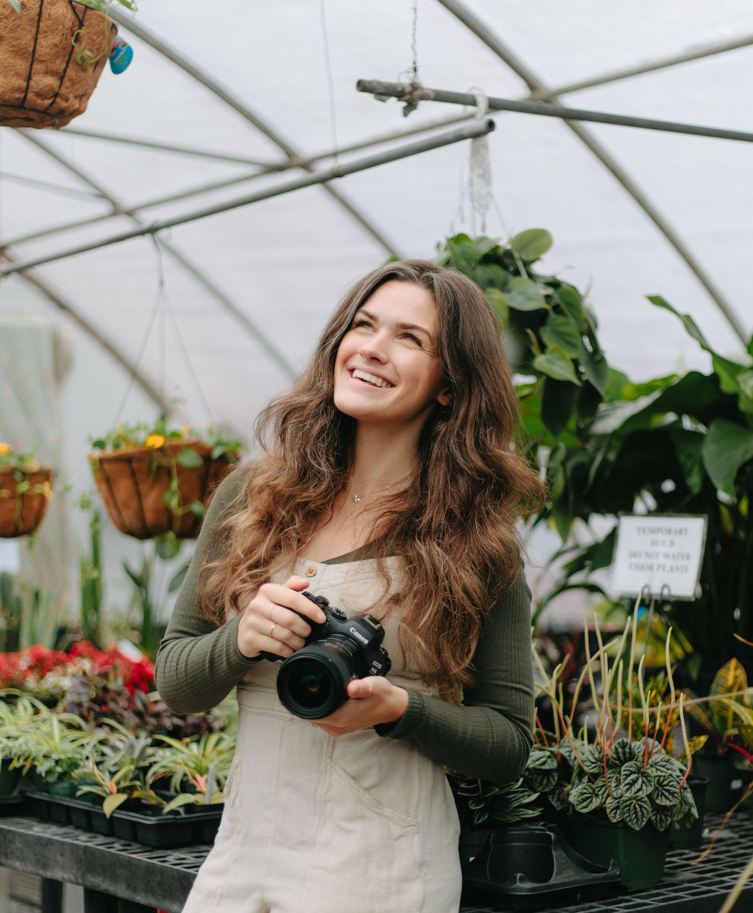 Person in green sweater and apron holds a camera while working in a greenhouse filled with plants.