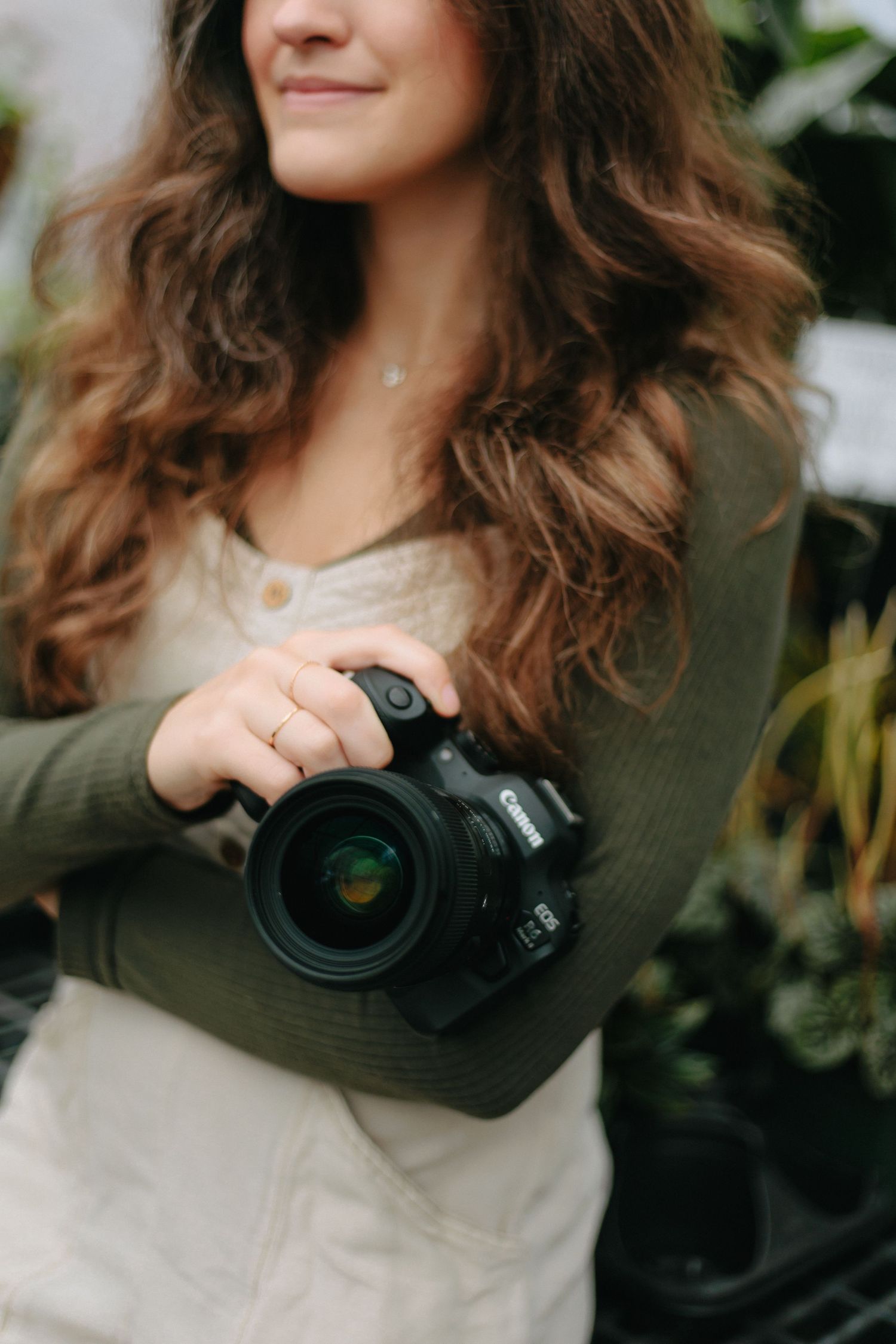 Photographer wearing olive green cardigan and white dress holds professional DSLR camera with large lens outdoors.