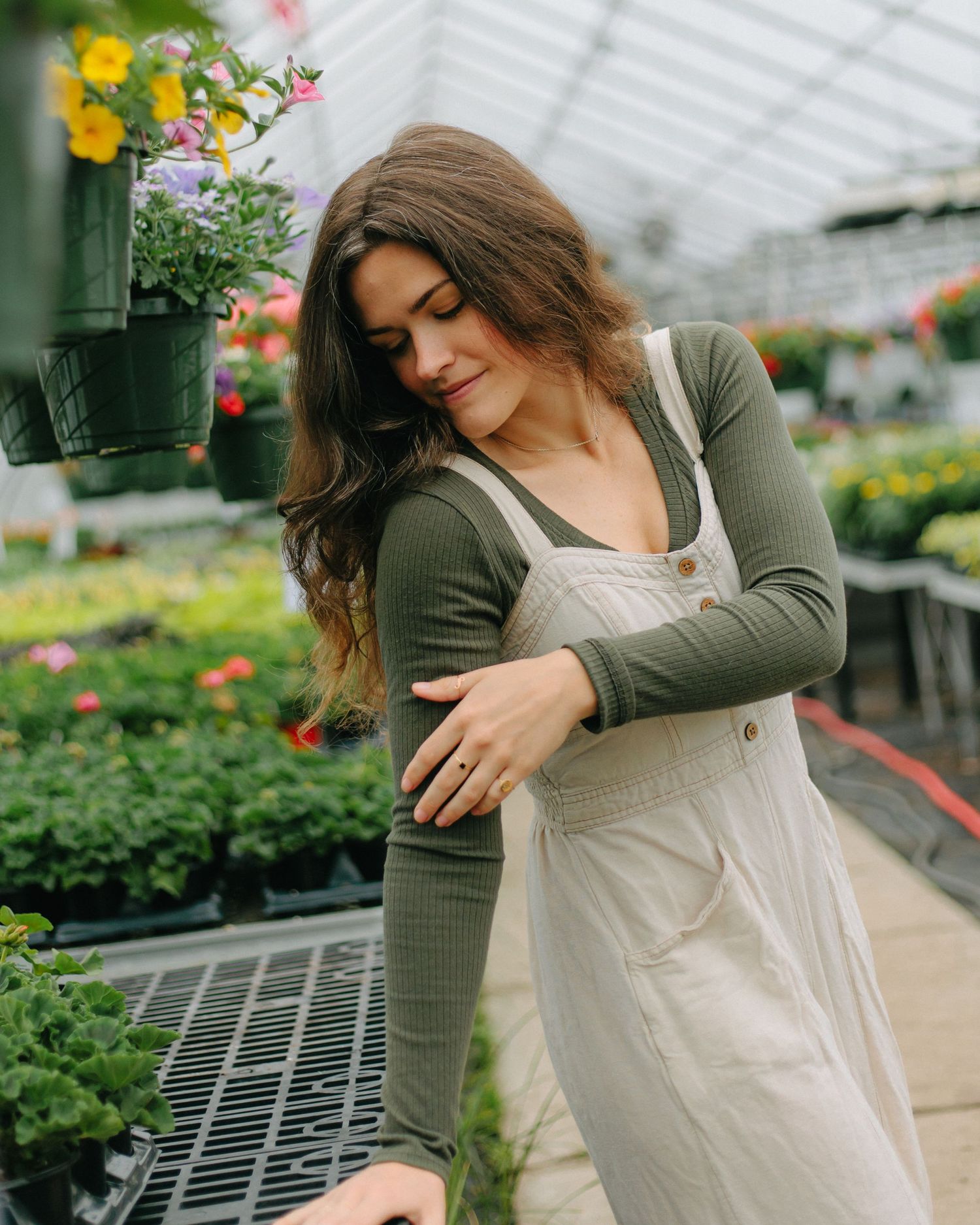 Someone wearing an olive cardigan and beige overalls tends to plants in a sunny greenhouse nursery filled with colorful flowers.