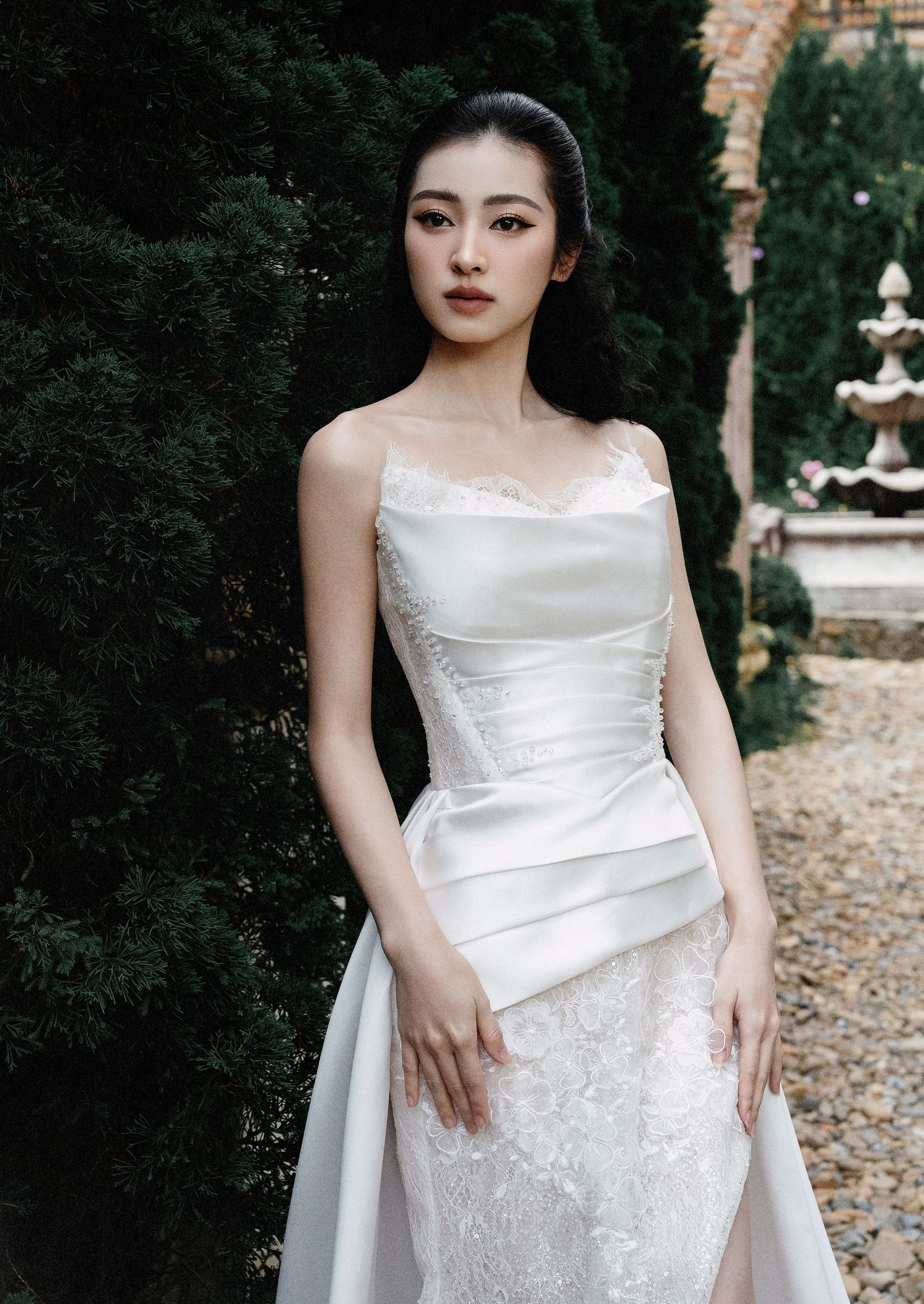 Elegant white wedding dress with intricate lace pattern and asymmetric draped detail photographed in garden setting.