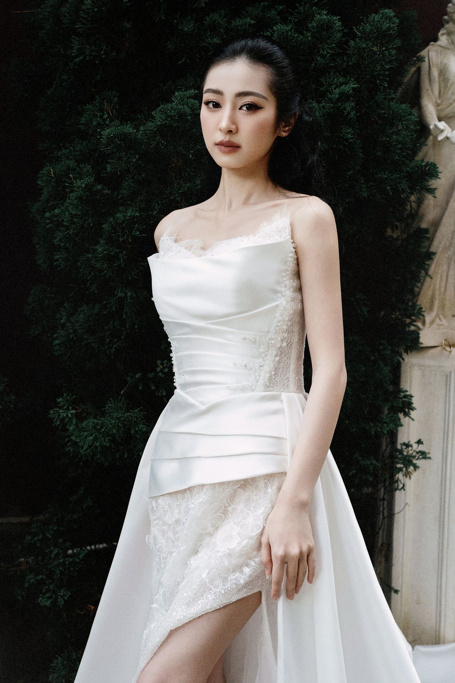 Elegant wedding dress with delicate lace details and spaghetti straps against a dark green foliage backdrop.