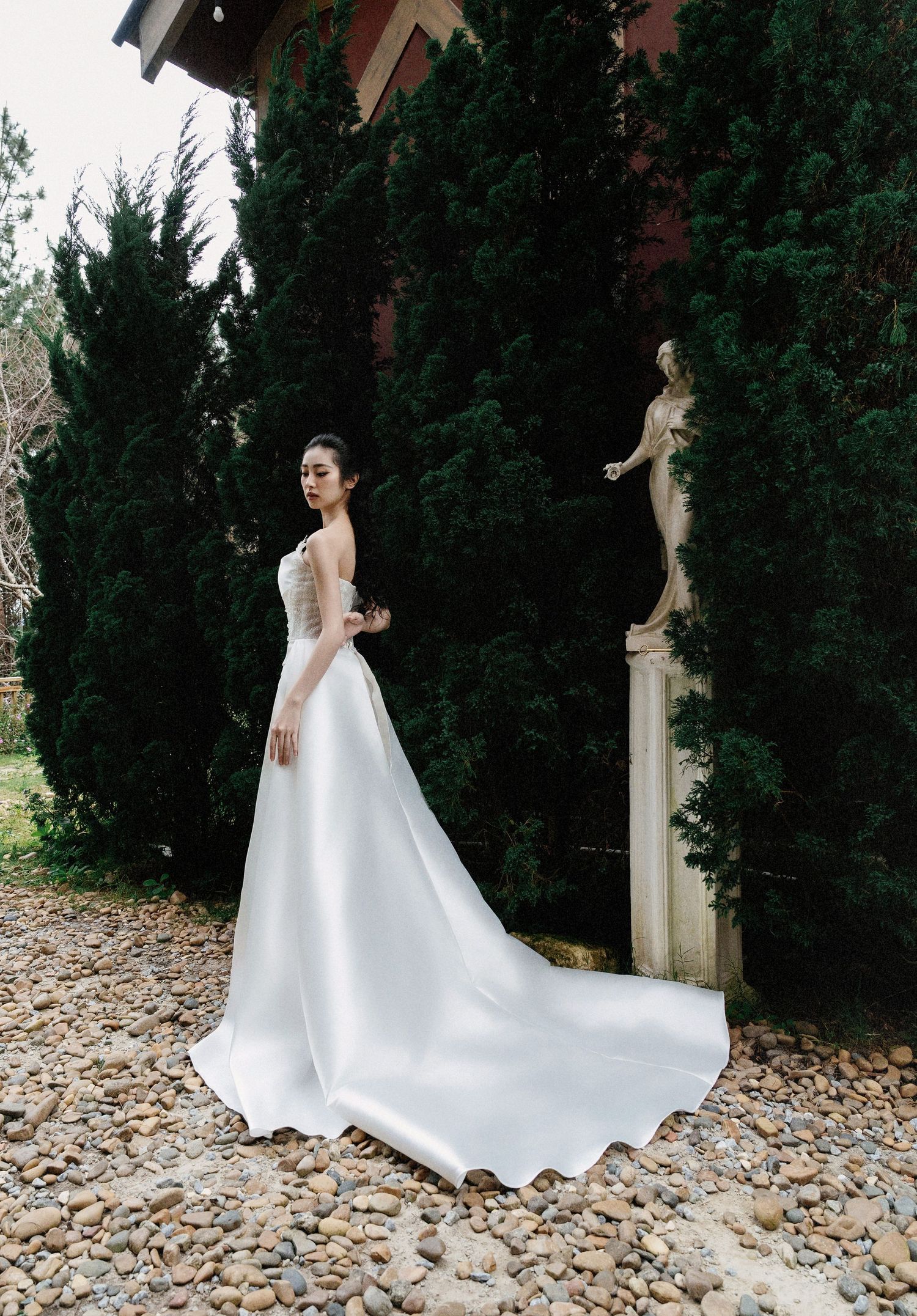 Elegant bride in flowing white wedding gown with long train poses in garden setting with classical statue and evergreen trees.