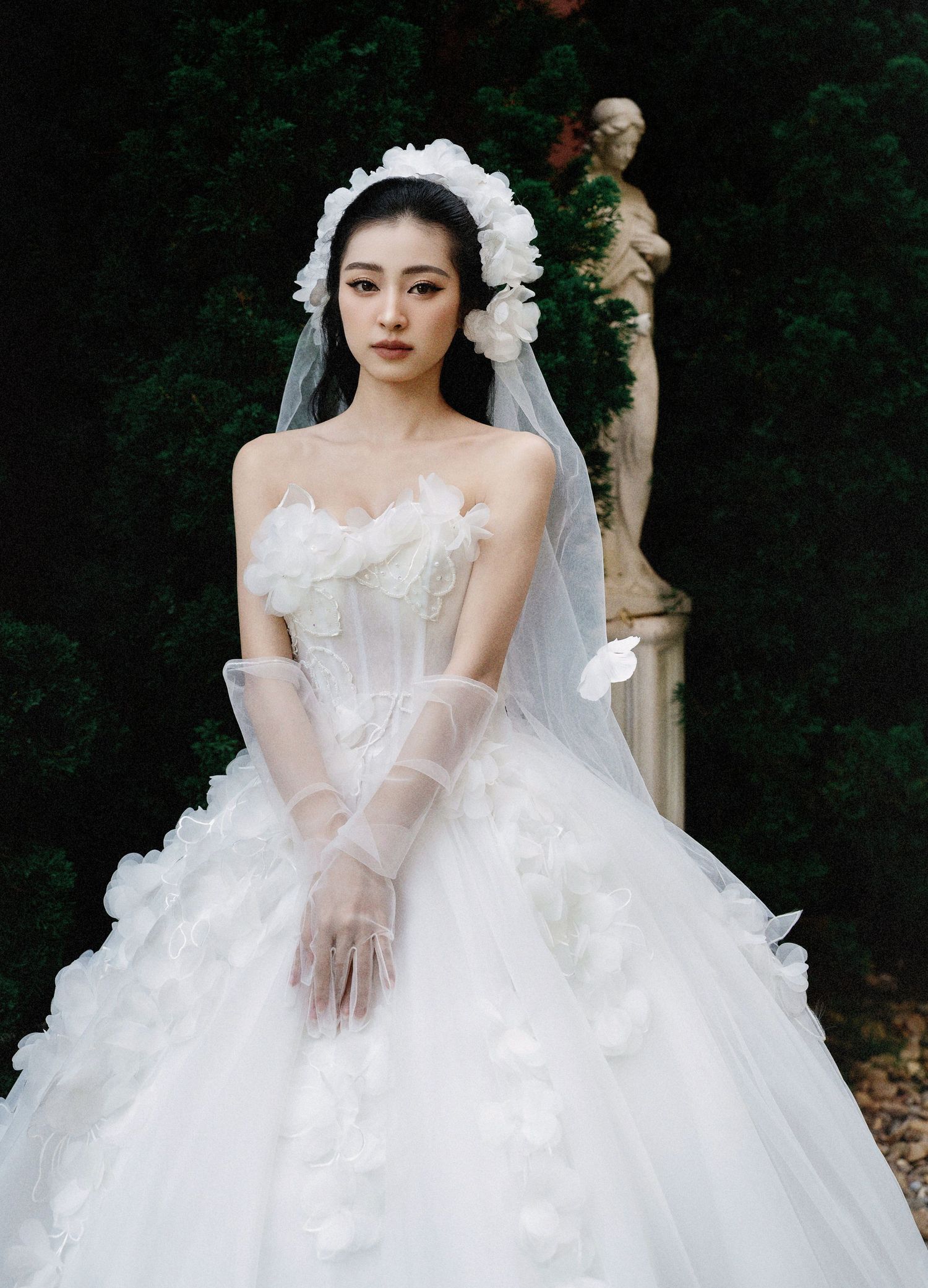 A bride in an elegant white ball gown wedding dress with tulle details and long gloves poses in front of a garden statue.