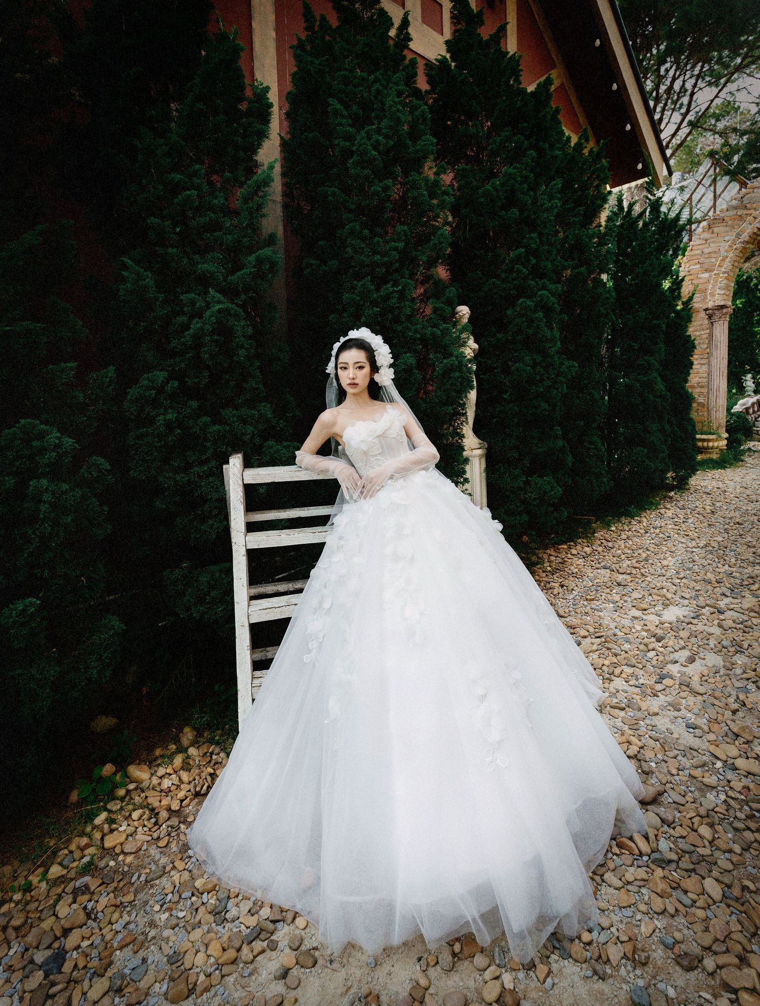 A stunning white ballgown wedding dress with layers of tulle photographed against evergreen trees on a garden path.