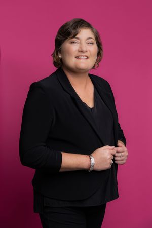 Professional headshot against a pink background showing a person in black business attire with a warm smile.