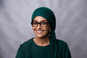 A person wearing glasses and dark green headwear smiles warmly against a gray studio backdrop.