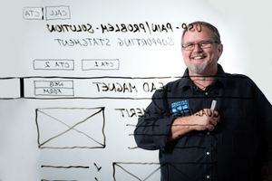 A person in a black shirt stands smiling in front of a whiteboard with sketched diagrams and user interface mockups.