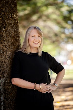 A person in a black top stands beside a tree trunk in natural outdoor lighting with a blurred background.