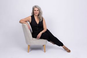 A person in a black outfit reclines elegantly on a light gray armchair against a white backdrop.