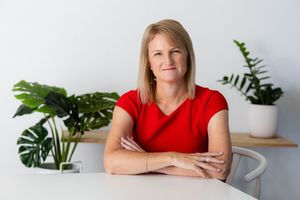 Professional portrait of someone in a red top sitting at a white desk with indoor plants in the background.