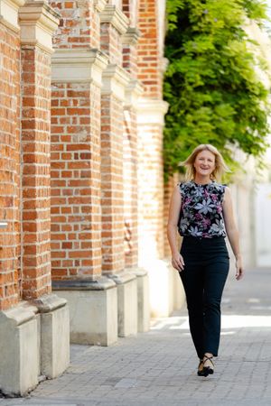 A person in a floral top and black pants strolling along a brick building on a paved walkway.