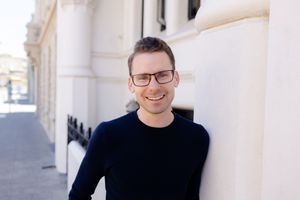 A person in glasses and a black sweater smiles while standing against a white building exterior.