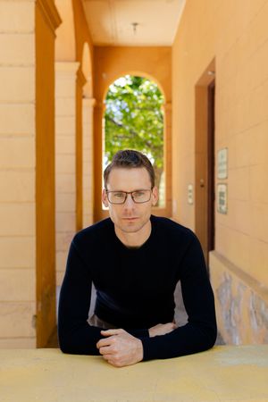 Person wearing glasses and black sweater sits in archway corridor with yellow walls and natural lighting.