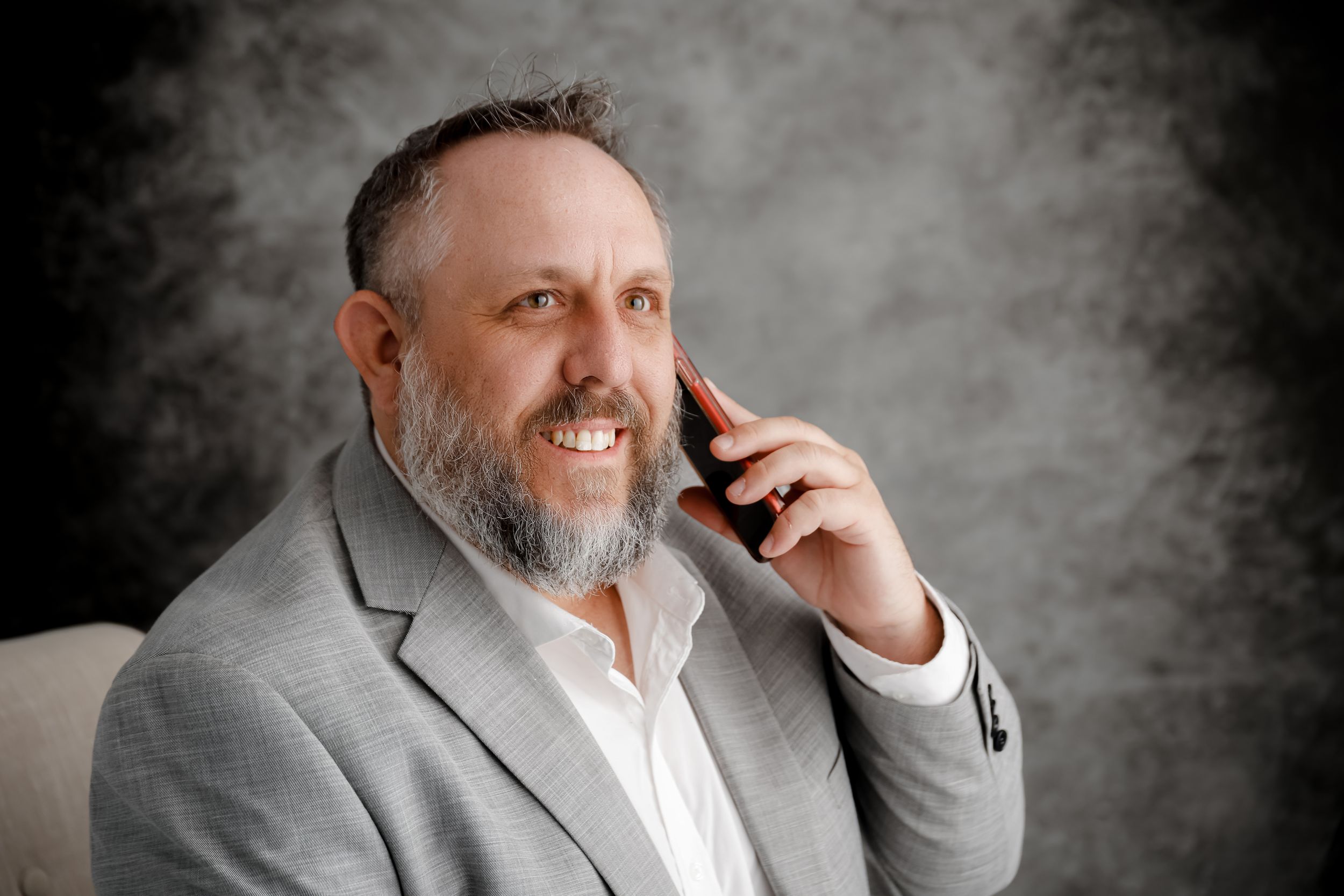 Professional portrait of a bearded businessman in a gray suit talking on a mobile phone against a gray background.
