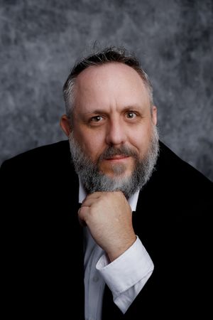 Professional studio portrait of a person with a salt-and-pepper beard wearing a black suit and white shirt in contemplative pose.