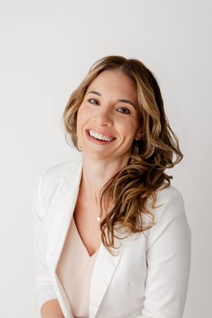 Professional headshot portrait of someone wearing a crisp white blazer and blush top while smiling warmly against light background.
