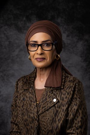 Professional portrait of a mature woman wearing glasses, a brown headscarf and animal print jacket against a dark background.