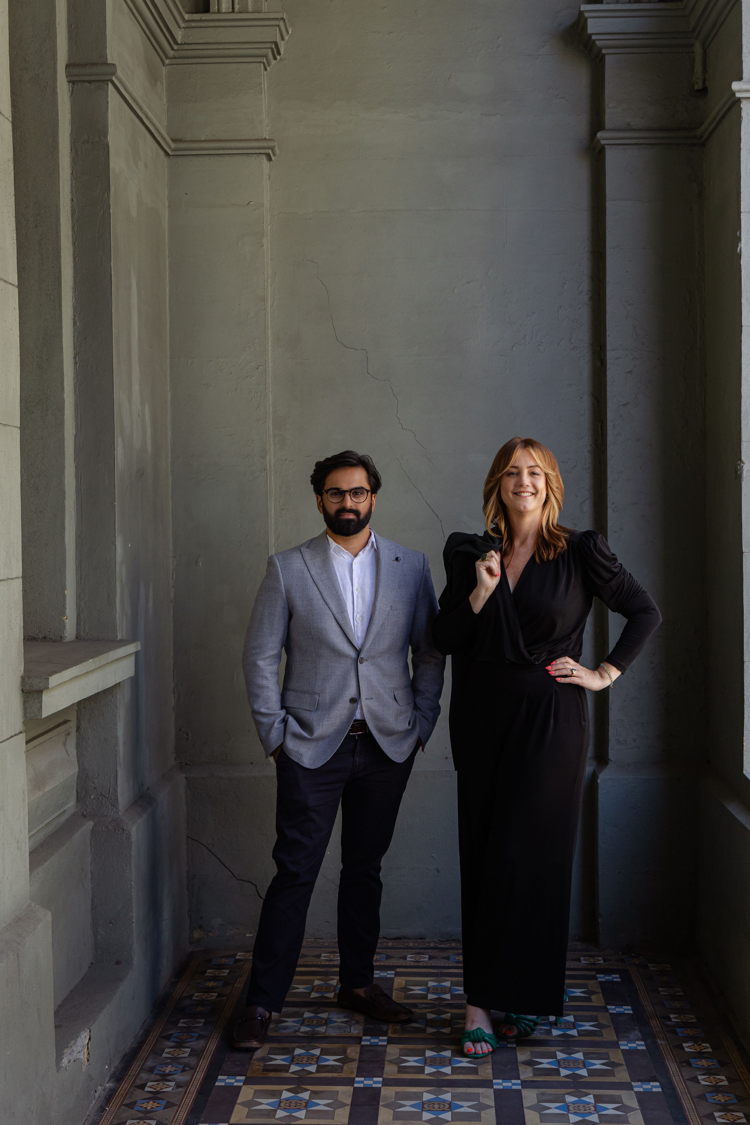 Two professionals in formal attire stand in an elegant hallway with ornate tiled floors and architectural columns.