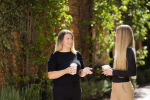 Two women in casual business attire holding coffee cups while having a conversation outdoors near brick wall and greenery.