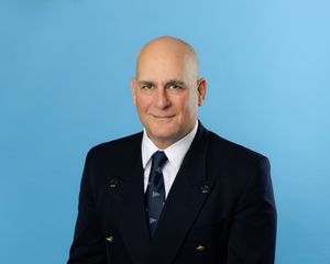 Professional headshot of someone in a dark business suit and blue tie against a bright blue background.
