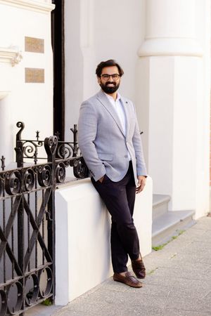 Professional portrait of a person in a light gray suit jacket and dark pants standing against a white building with ornate railings.
