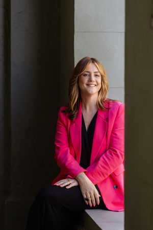 Professional portrait of someone in a vibrant pink blazer and black top leaning against a column in natural lighting.