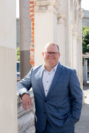 Professional in a blue checkered suit and glasses stands casually by a white pillar outdoors in natural lighting.
