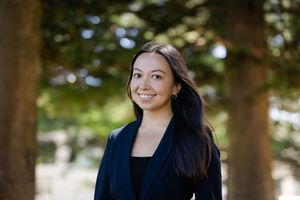 Professional portrait of a smiling person in a dark blazer outdoors with trees in the soft-focus background.