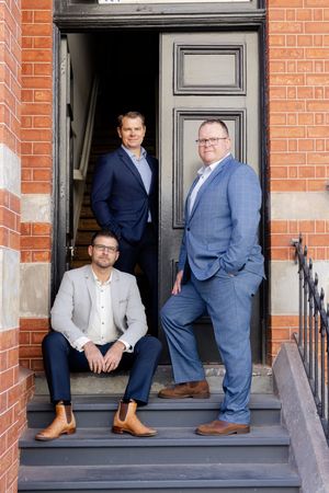 Three business professionals in suits pose together on stone steps of a brick building with a black doorway.