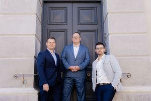 Three business professionals in suits pose together in front of a large gray door with a classic architectural style.