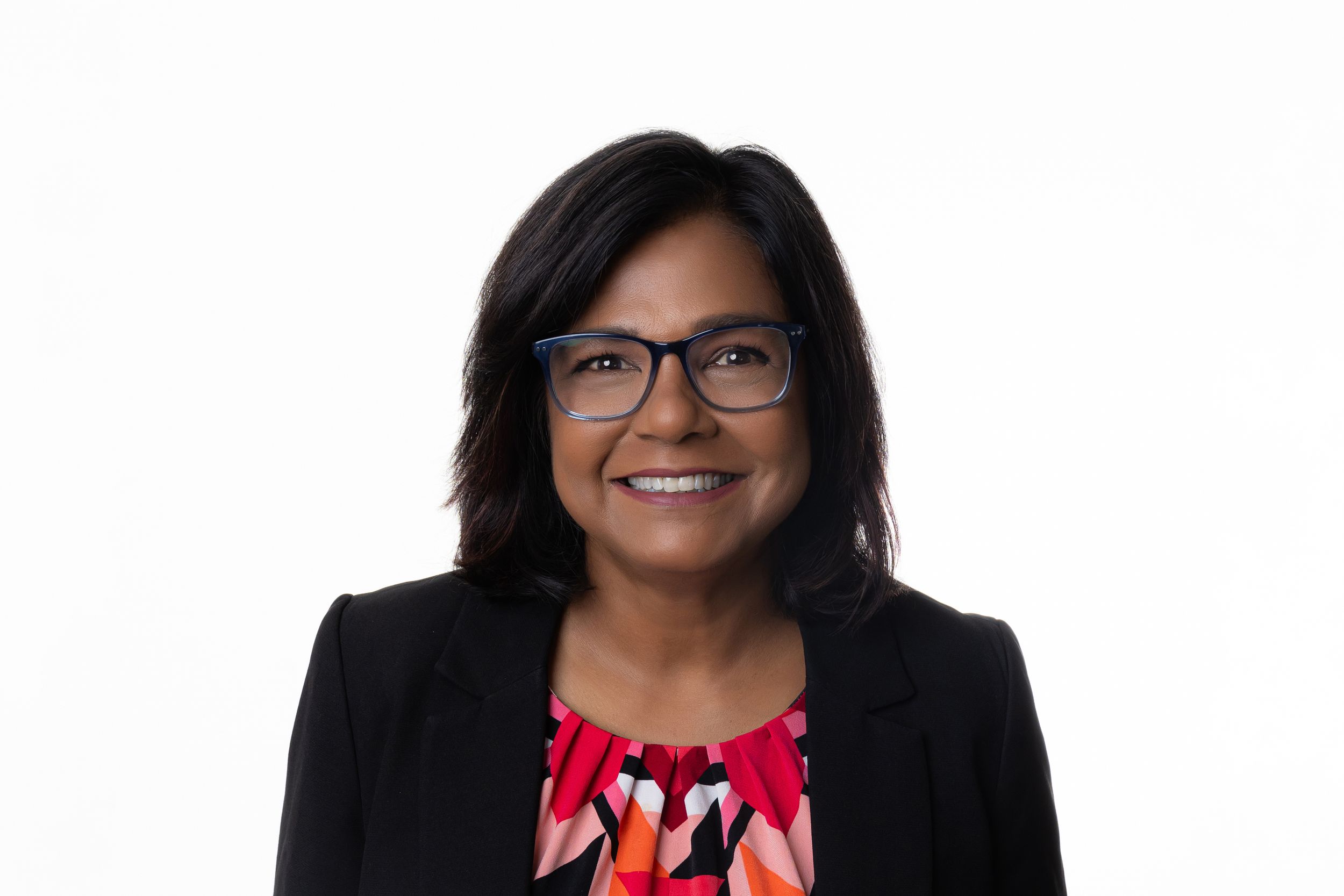 Professional headshot of someone wearing glasses, a black blazer and patterned red top against white background.