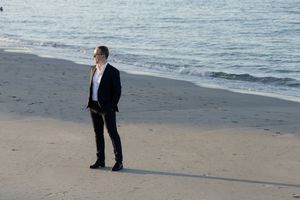 A person in a dark business suit stands alone on a sandy beach at the water's edge during daytime.