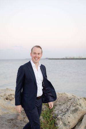 Person in navy suit and white shirt smiling while standing on rocky coastal shore at sunset with water in background.