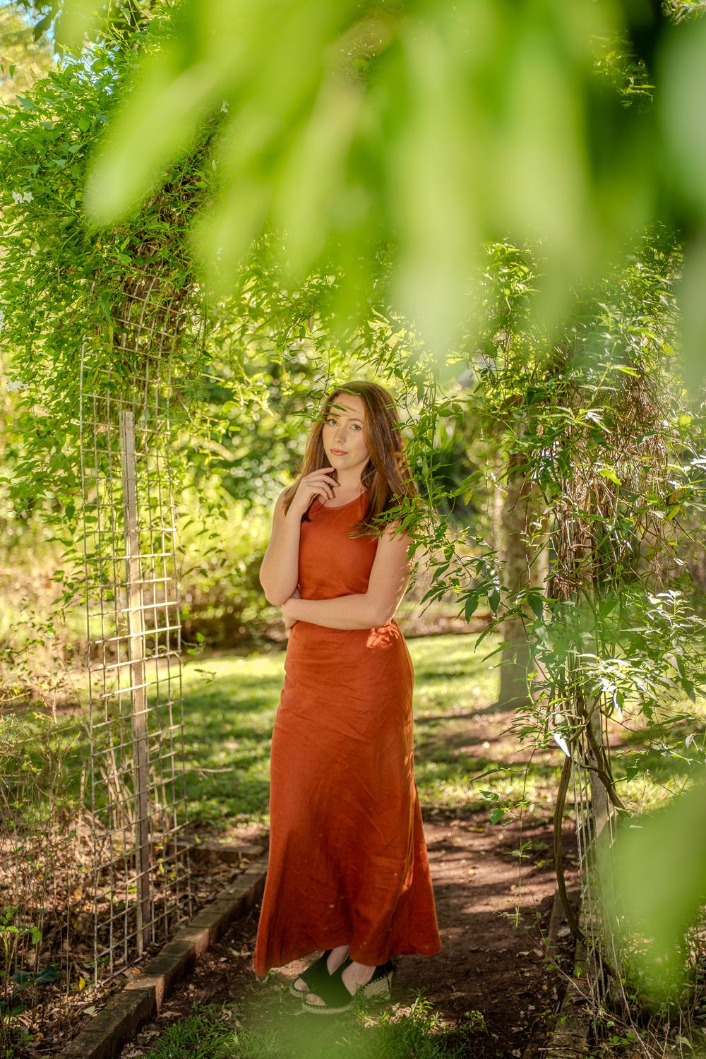 A person in a rust-colored maxi dress stands thoughtfully on a garden path surrounded by lush greenery and sunlight.