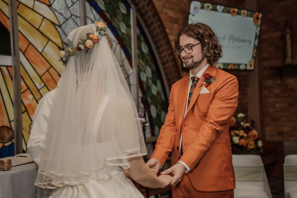 Person in a bright orange suit holds papers during an intimate wedding ceremony with a veiled figure in white.